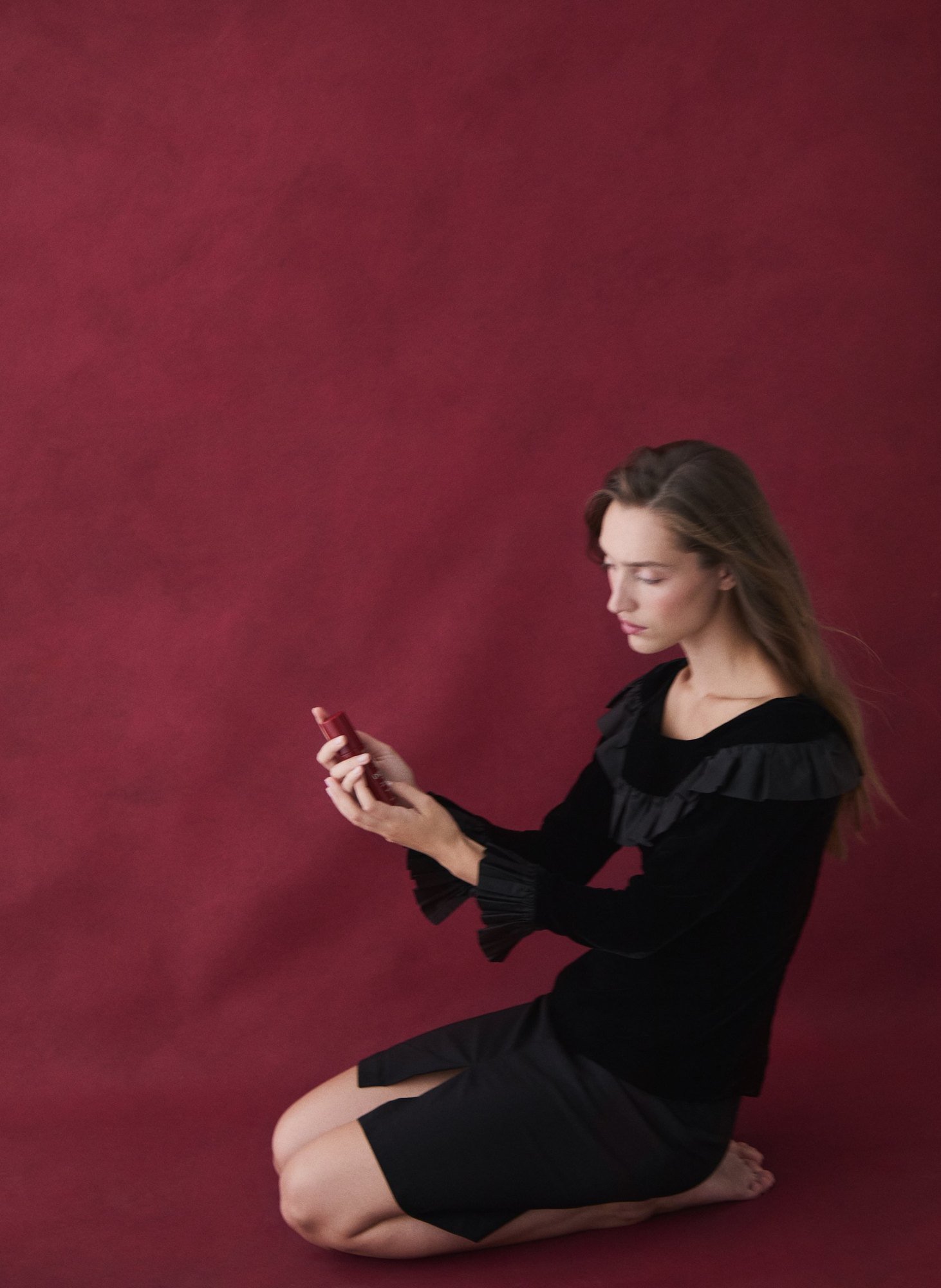 Kneeling against a burgundy backdrop in black velvet, long straight hair, serum in hand