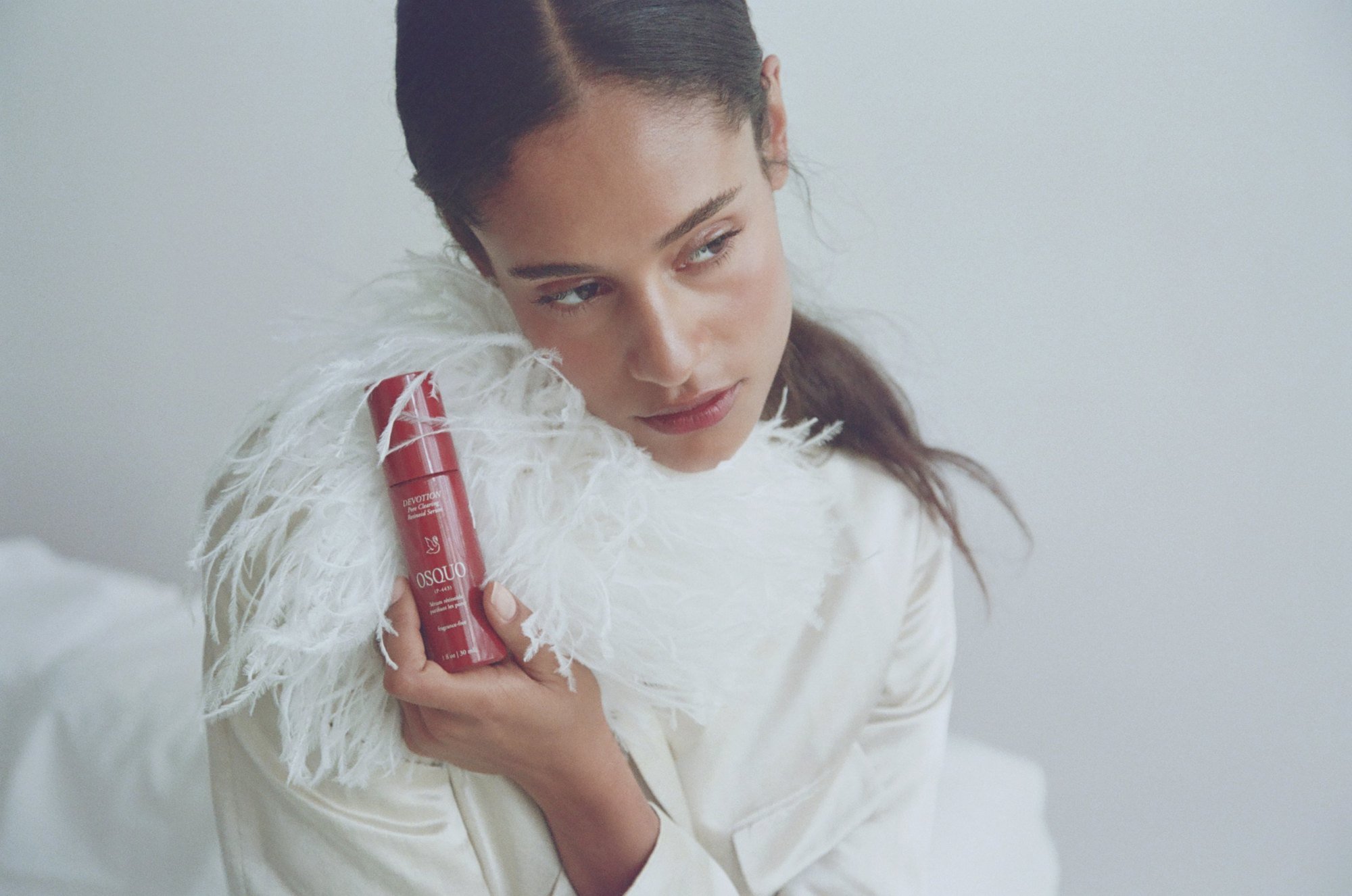 White feather jacket, sleek center part pulled back, holding the red serum