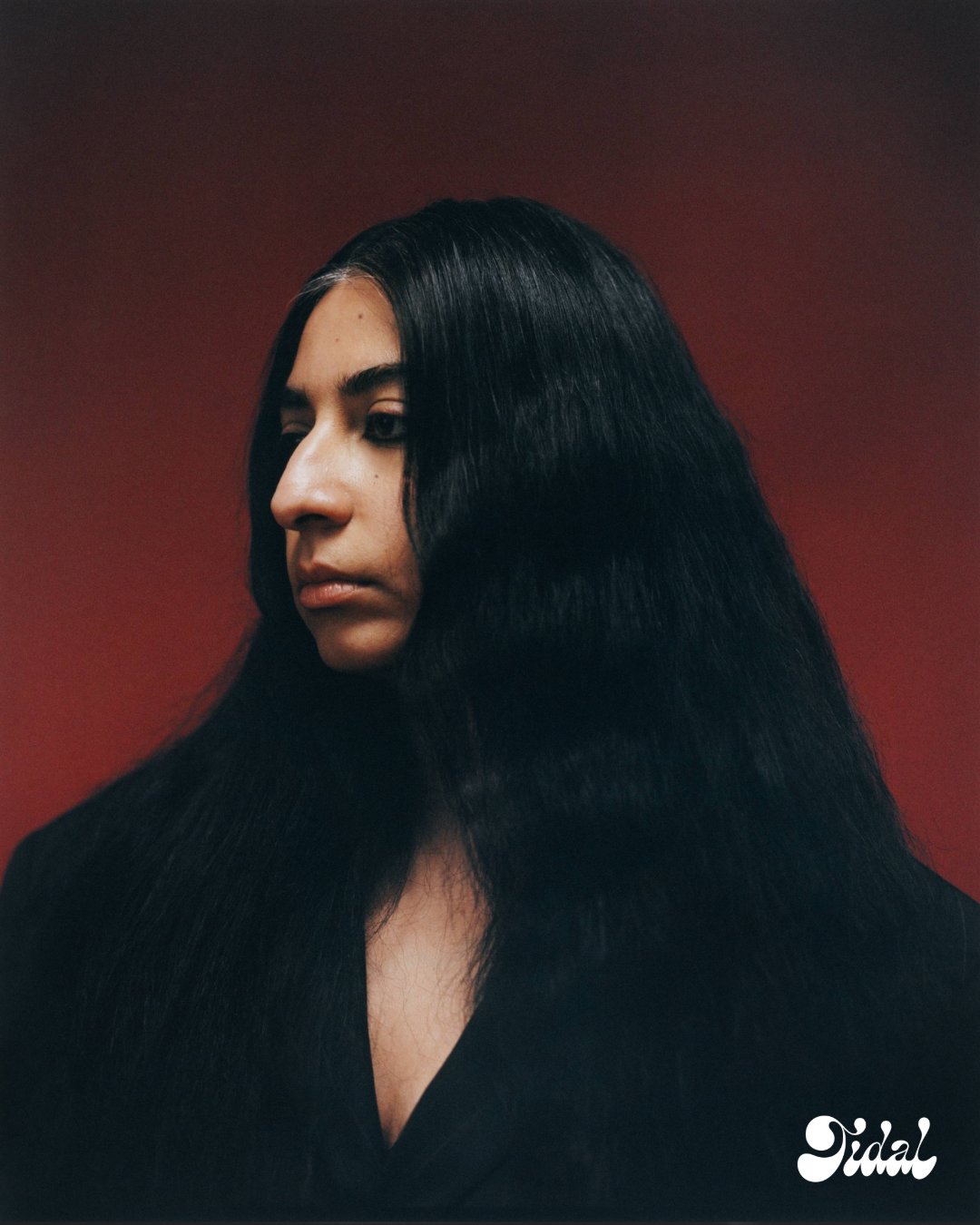 Arooj Aftab in profile against a deep red backdrop, long black hair worn loose and glossy